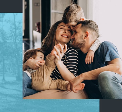 Eine vierköpfige Familie sitzt eng beieinander, hält Händchen und umarmt sich in einer gemütlichen Inneneinrichtung.
