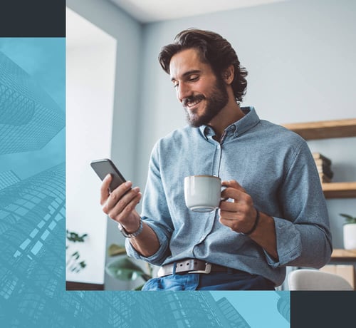 Ein Mann in einem legeren Hemd, der in der einen Hand eine Kaffeetasse und in der anderen ein Smartphone hält und drinnen in der Nähe eines Fensters und einiger Regale sitzt.