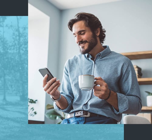 Ein Mann in einem legeren Hemd, der in der einen Hand eine Kaffeetasse und in der anderen ein Smartphone hält und drinnen in der Nähe eines Fensters und einiger Regale sitzt.