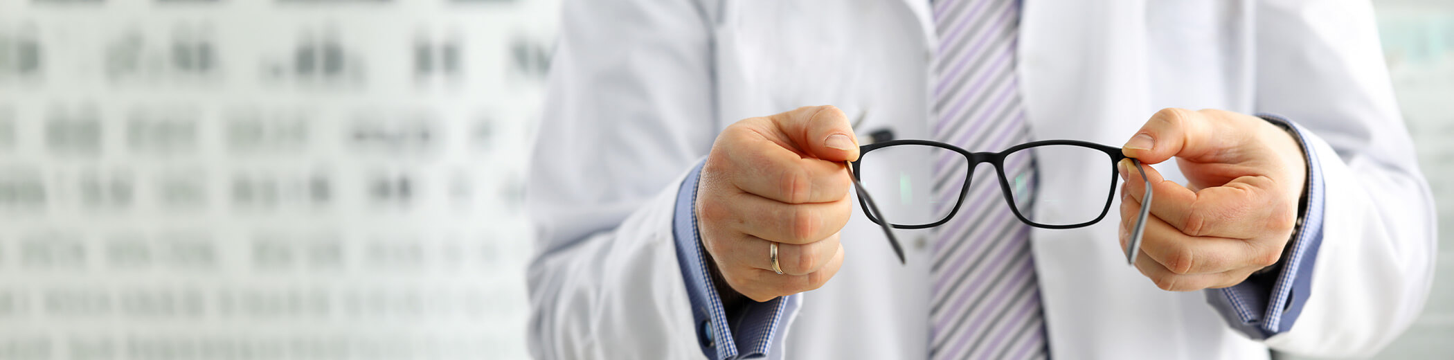 Ein Optiker hält eine Brille in der Hand, im Hintergrund ist eine unscharfe Sehtafel zu sehen