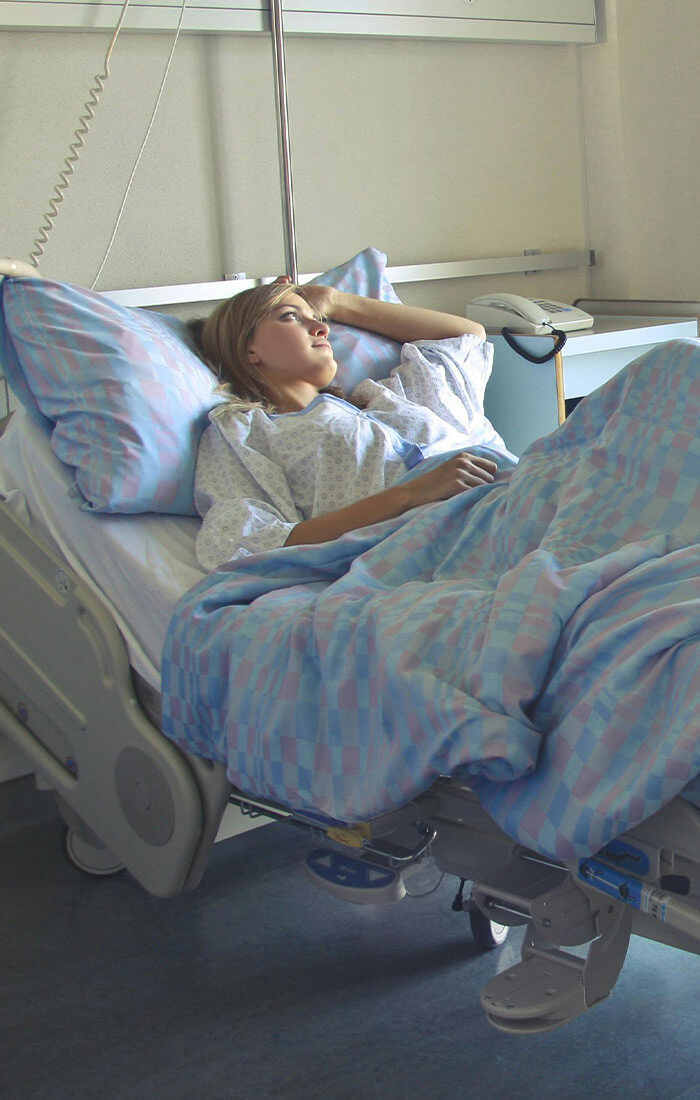 eine Patientin liegt in einem Krankenhausbett mit blau gemusterter Bettwäsche, ihr Blick geht Richtung Fenster