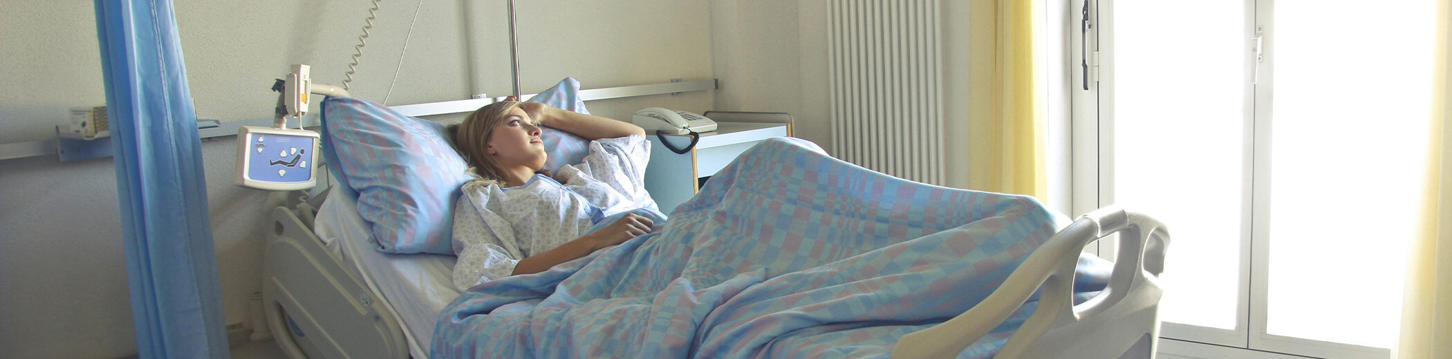 eine Patientin liegt in einem Krankenhausbett mit blau gemusterter Bettwäsche neben einem Fenster