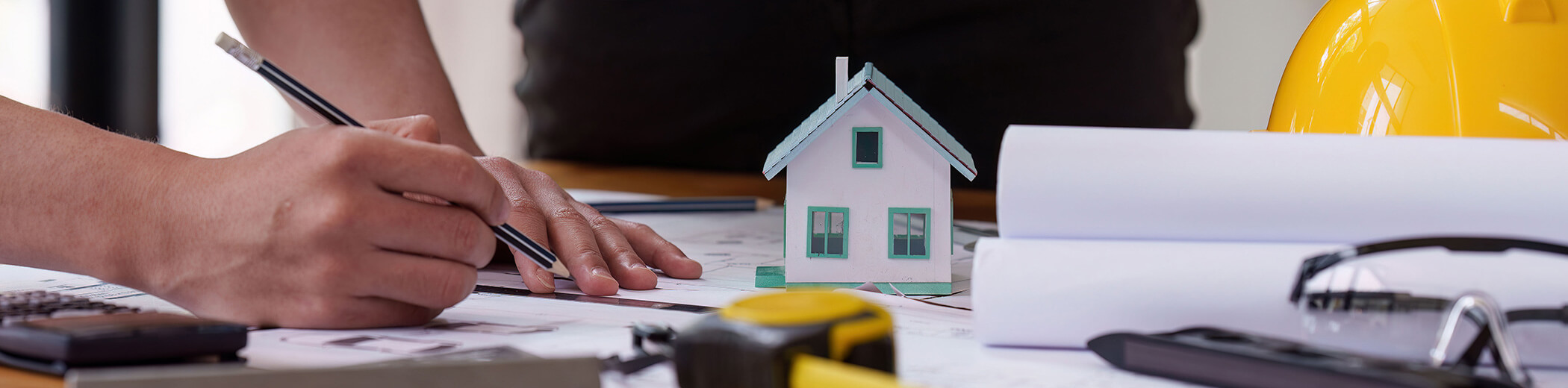 eine Person überprüft Architekturpläne mit einem Modellhaus und einem Bauhelm auf dem Schreibtisch