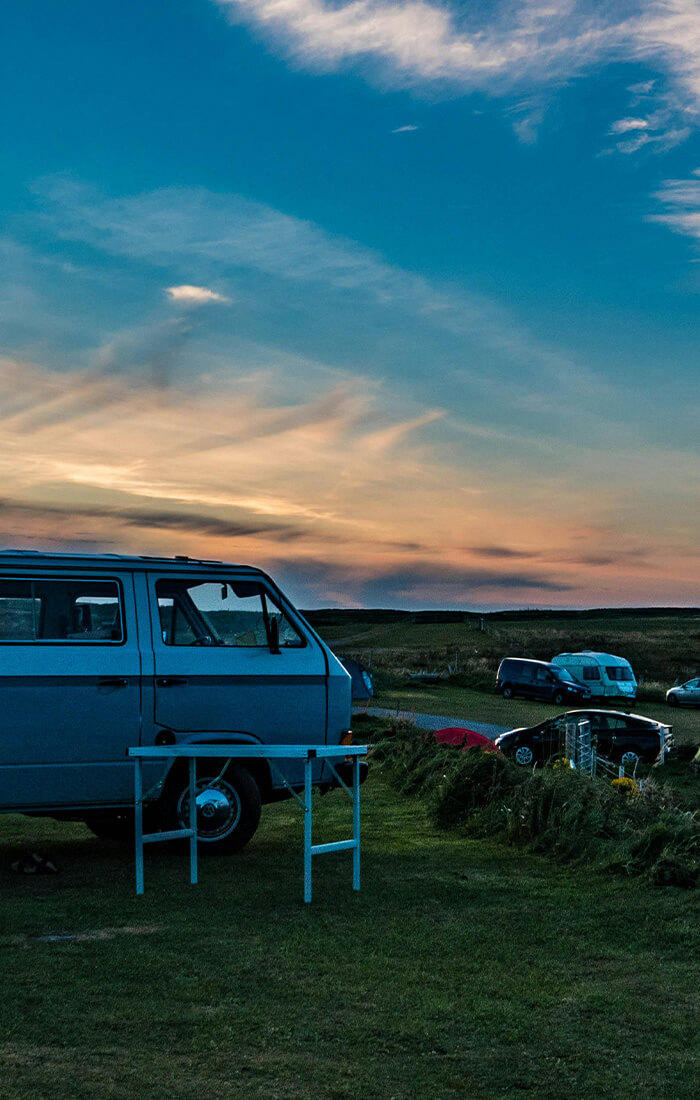 Ein Campingplatz bei Sonnenuntergang mit Wohnmobilen, Zelten und Autos uauf einer Wiese unter einem farbenprächtigen Himmel.