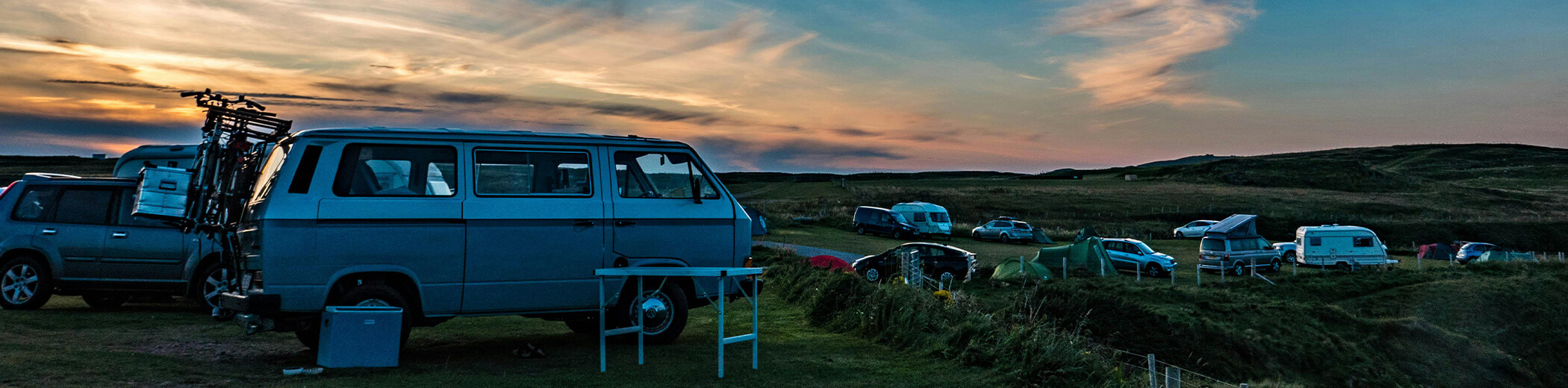 Ein Campingplatz bei Sonnenuntergang mit Wohnmobilen, Zelten und Autos uauf einer Wiese unter einem farbenprächtigen Himmel.