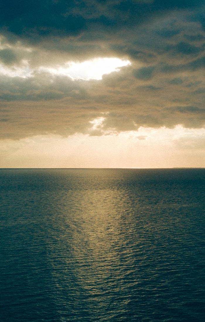 Ein ruhiger Ozean unter bewölktem Himmel, während am Horizont Sonnenstrahlen durch die Wolken brechen.