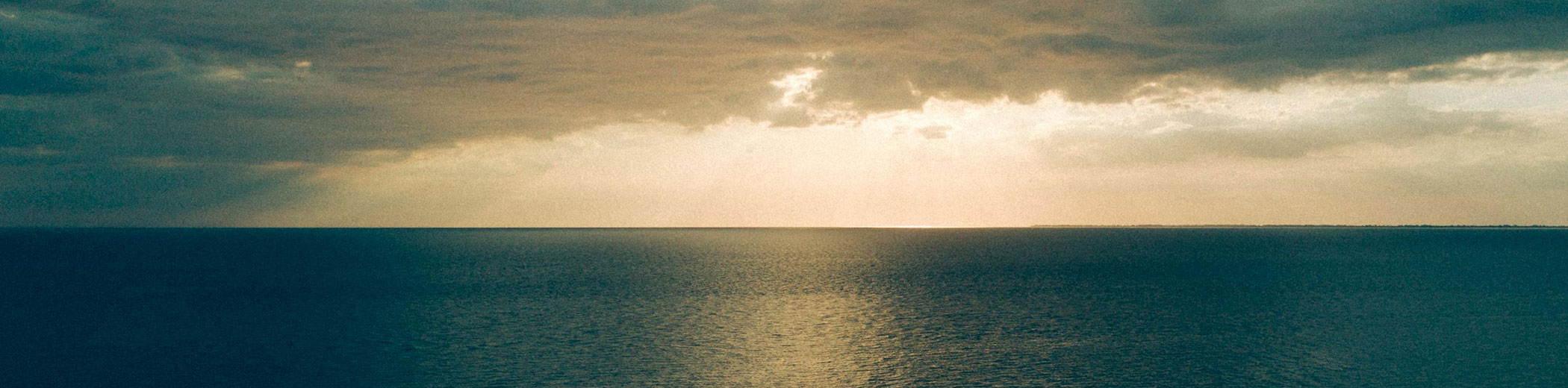 Ein ruhiger Ozean unter bewölktem Himmel, während am Horizont Sonnenstrahlen durch die Wolken brechen.