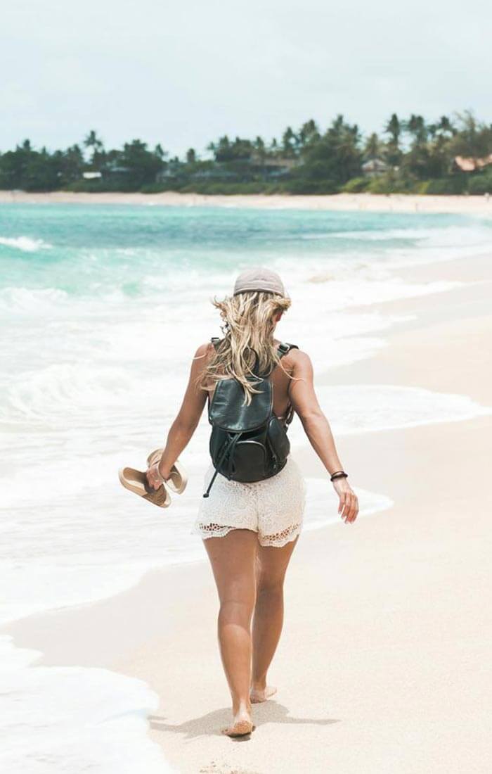 Eine Frau mit Hut und Rucksack, die barfuß an einem Sandstrand am türkisfarbenen Meer entlangläuft und ihre Sandalen in der Hand hält.