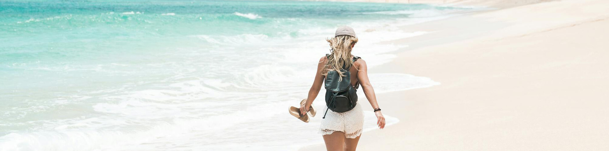 Eine Frau mit Hut und Rucksack, die barfuß an einem Sandstrand am türkisfarbenen Meer entlangläuft und ihre Sandalen in der Hand hält.