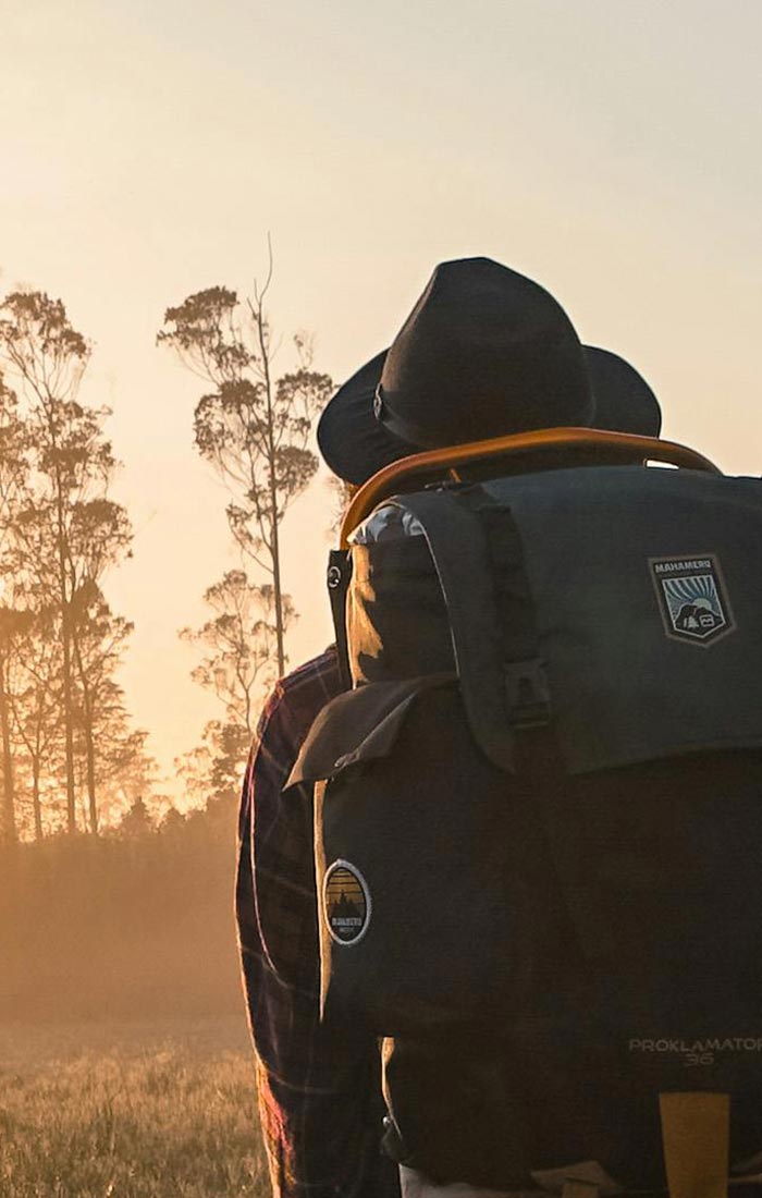 ein Mann mit Hut und Rucksack wandert in Richtung eines Waldes und des Sonnenaufgangs